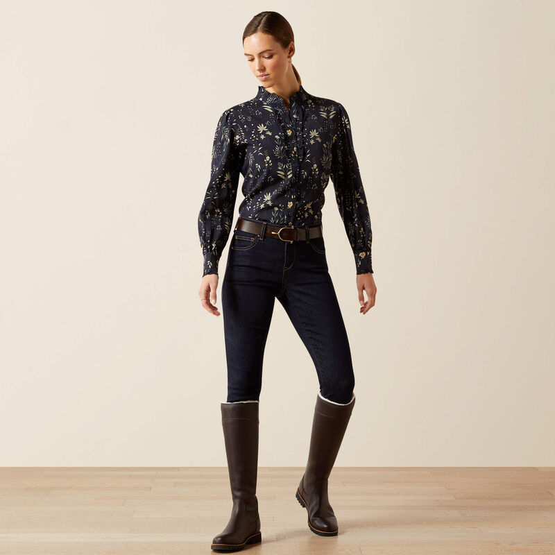 Woman wearing a patterned shirt, dark jeans, and brown riding boots on a plain background