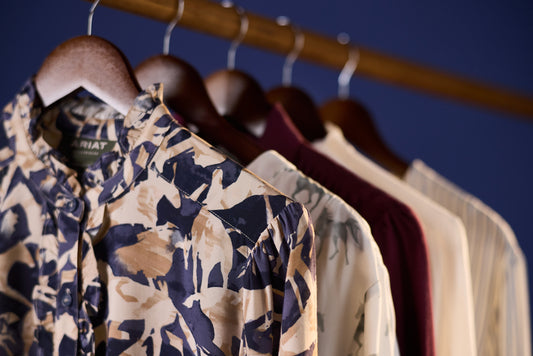 A close-up of a patterned button-up shirt hanging on a wooden hanger from a clothing rack, with other shirts in the background against a dark blue backdrop.