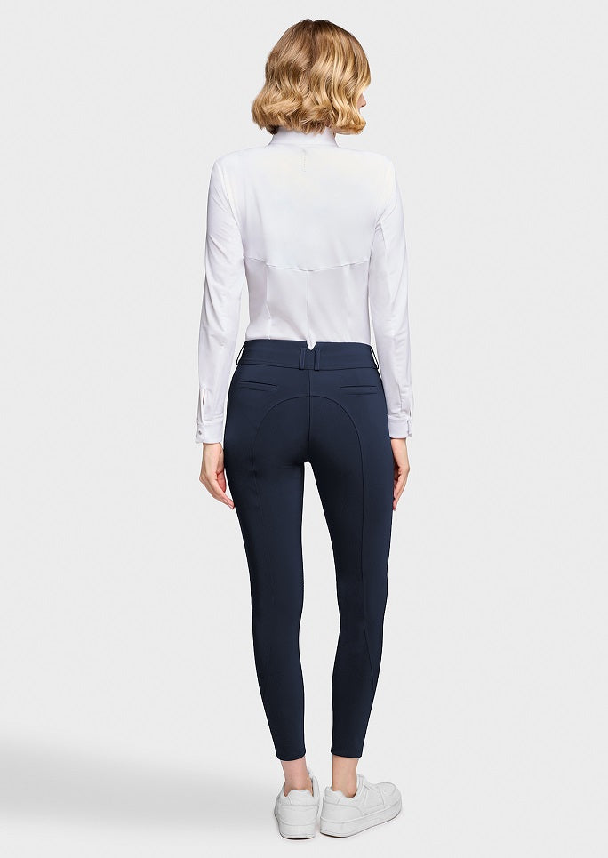 A woman viewed from the back wearing a white long-sleeve blouse, navy blue fitted pants, and white sneakers against a light gray studio background.