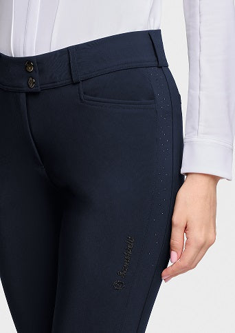 Close-up of a person wearing dark navy equestrian riding pants with a white shirt, showing the side pocket, button waistband, and a small embroidered brand logo near the lower leg.