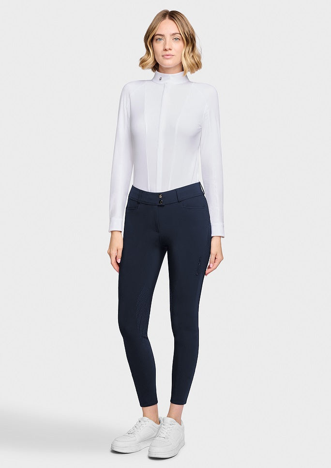 A woman modeling a white long-sleeve mock-neck shirt and navy blue riding breeches with white sneakers, set against a plain light gray studio background.
