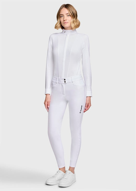 A woman modeling a white long-sleeve athletic zip-front top and matching white fitted pants with white sneakers, photographed against a light gray studio background.