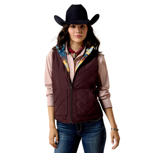 A woman modeling a dark burgundy quilted zip-up vest over a pink striped shirt, accessorized with a black cowboy hat, red neckerchief, and blue jeans, against a plain white background.