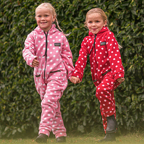 Two children in pink and red polka dot onesies holding hands in front of a green hedge.