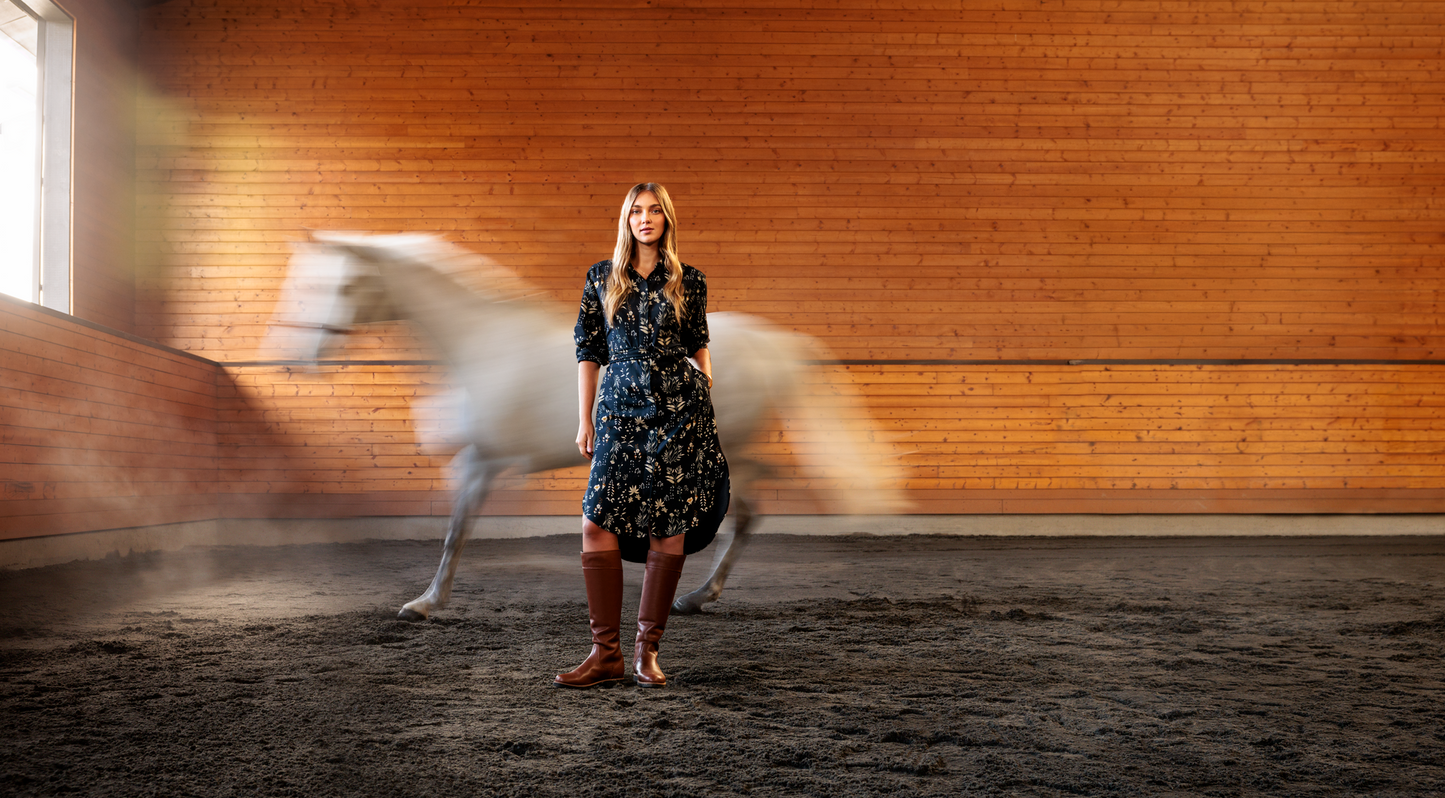 Woman in a floral dress standing in an indoor equestrian arena with a horse in the background.