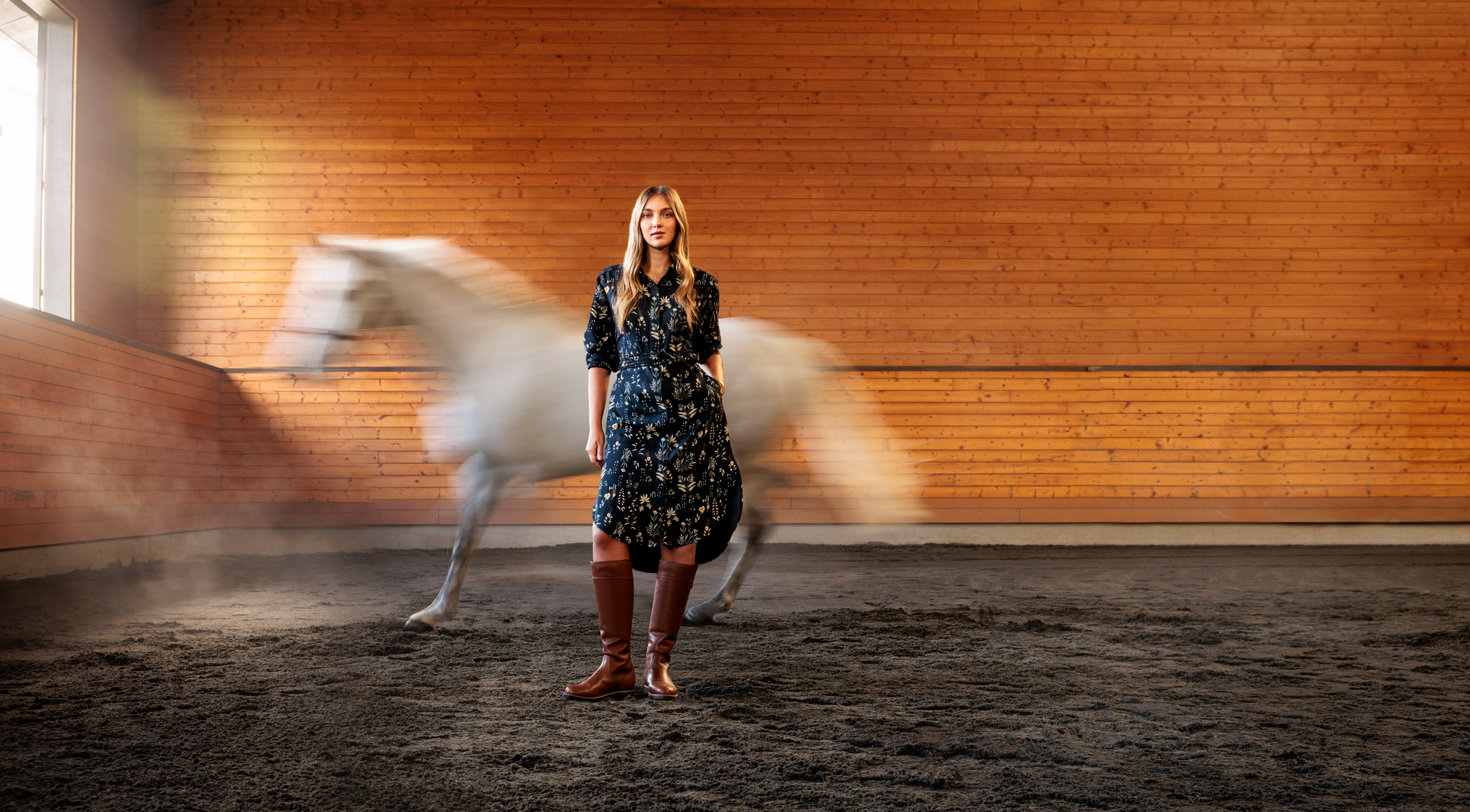 Woman in a floral dress standing in an indoor equestrian arena with a horse in the background.
