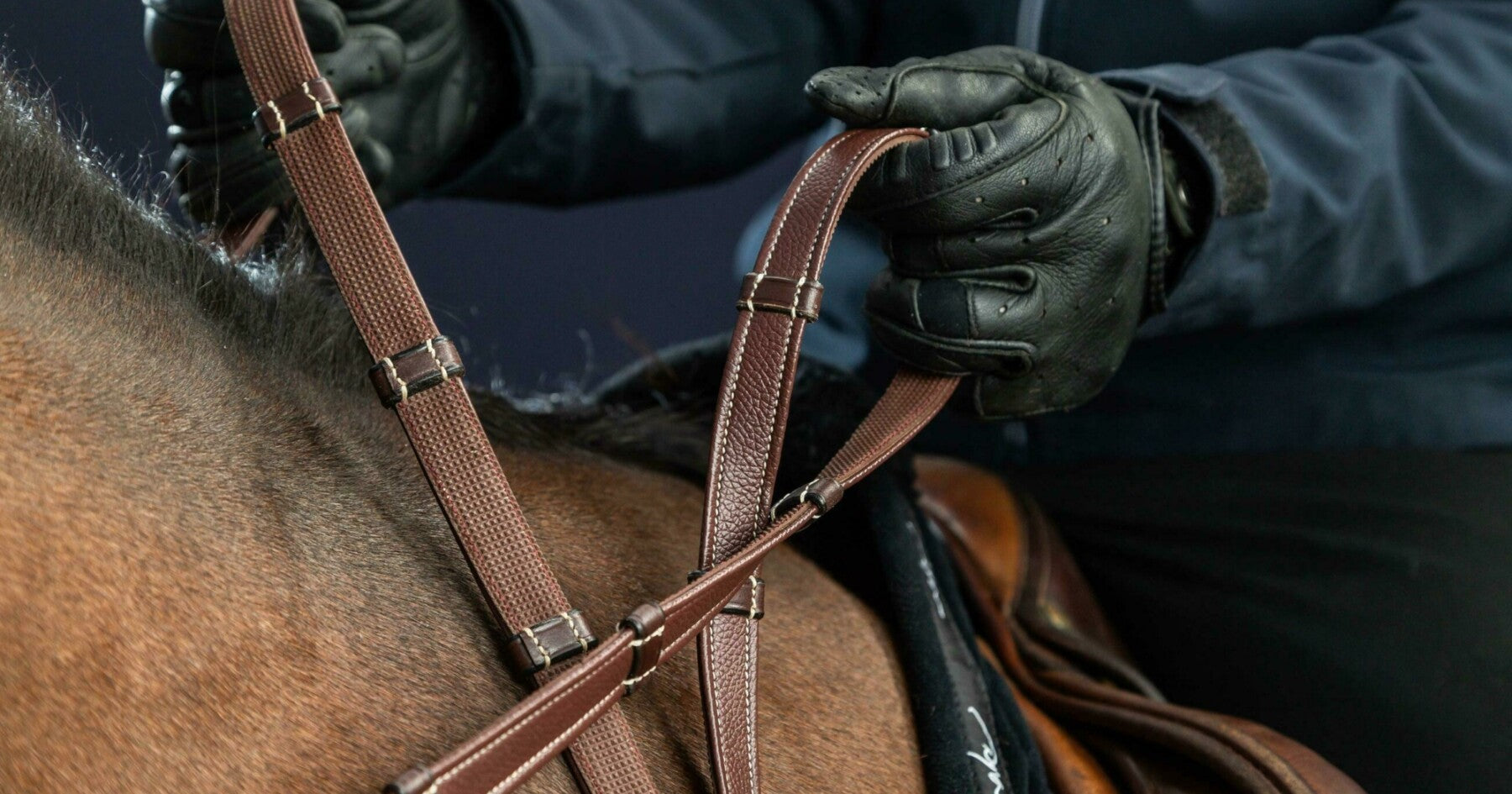 Person adjusting a brown bridle on a horse's head, wearing dark gloves against a dark background.