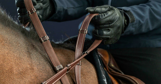 Person adjusting a brown bridle on a horse's head, wearing dark gloves against a dark background.