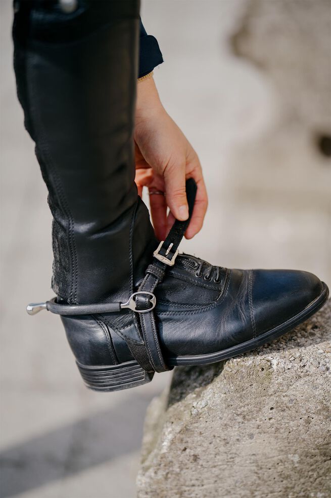 Person adjusting a black leather boot with a strap on a concrete surface