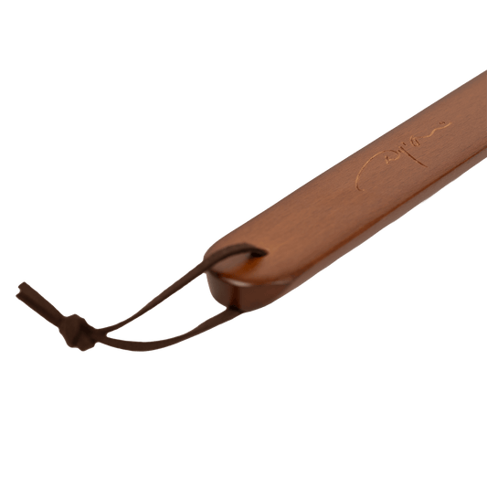 A close-up of a brown wooden paddle or handle with a dark brown leather loop and cord, featuring an engraved script logo, set against a plain white background.