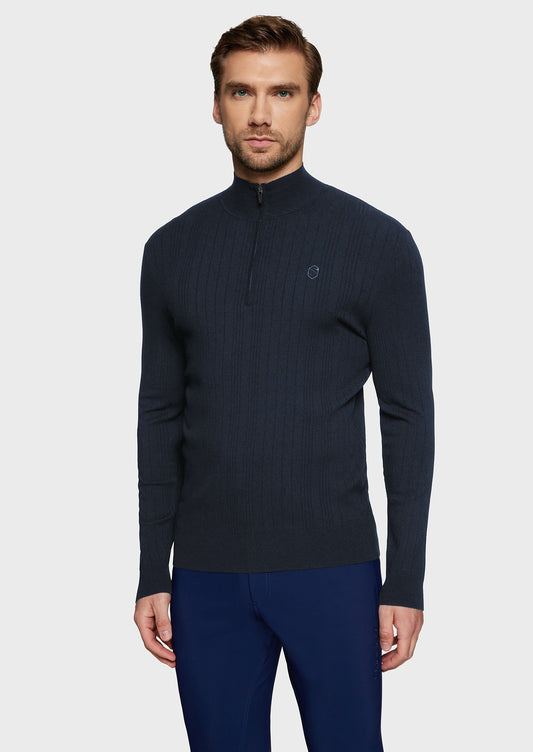 Man wearing a navy knitted jumper and blue breeches on a beige background