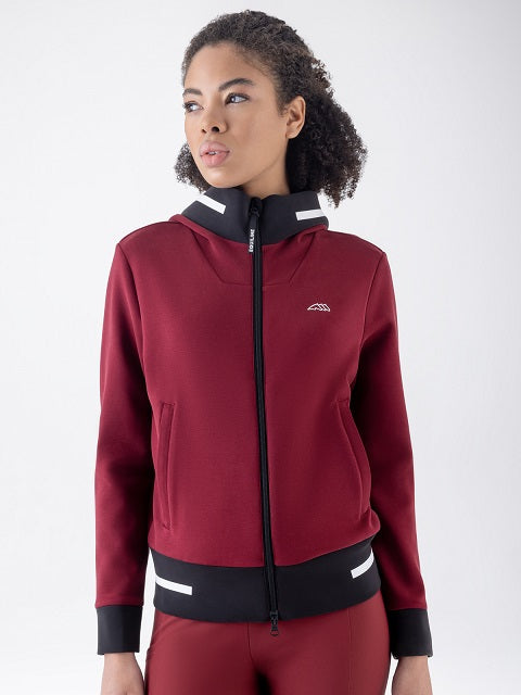 A woman wearing a burgundy zip-up hooded jacket with black and white trim, paired with matching burgundy athletic pants, photographed against a light gray studio background.