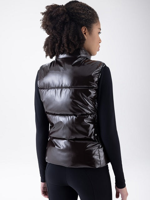 A woman shown from the back wearing a shiny black puffer vest over a black long-sleeve top and black leggings, photographed against a light gray studio background.