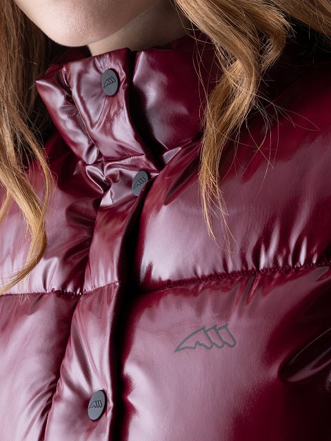 Close-up of a shiny burgundy puffer jacket with a high collar, dark buttons, and a small gray stylized logo on the chest, worn by a model with light brown hair.