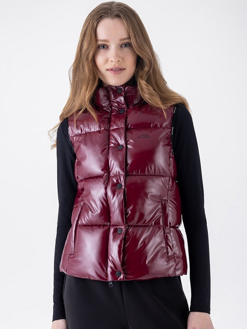 A woman wearing a shiny burgundy puffer vest over a black long-sleeve top, photographed against a plain light gray studio background.
