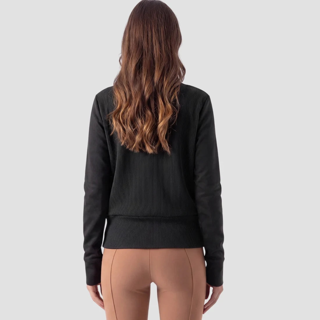 A woman viewed from the back wearing a black ribbed long-sleeve top and tan leggings against a light gray studio background.