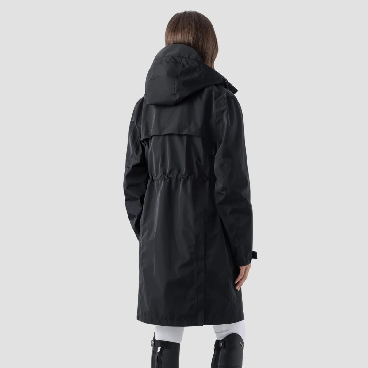 A woman modeling a black hooded raincoat against a light gray studio background. The coat has a small white logo on the left chest and a white logo on the sleeve.