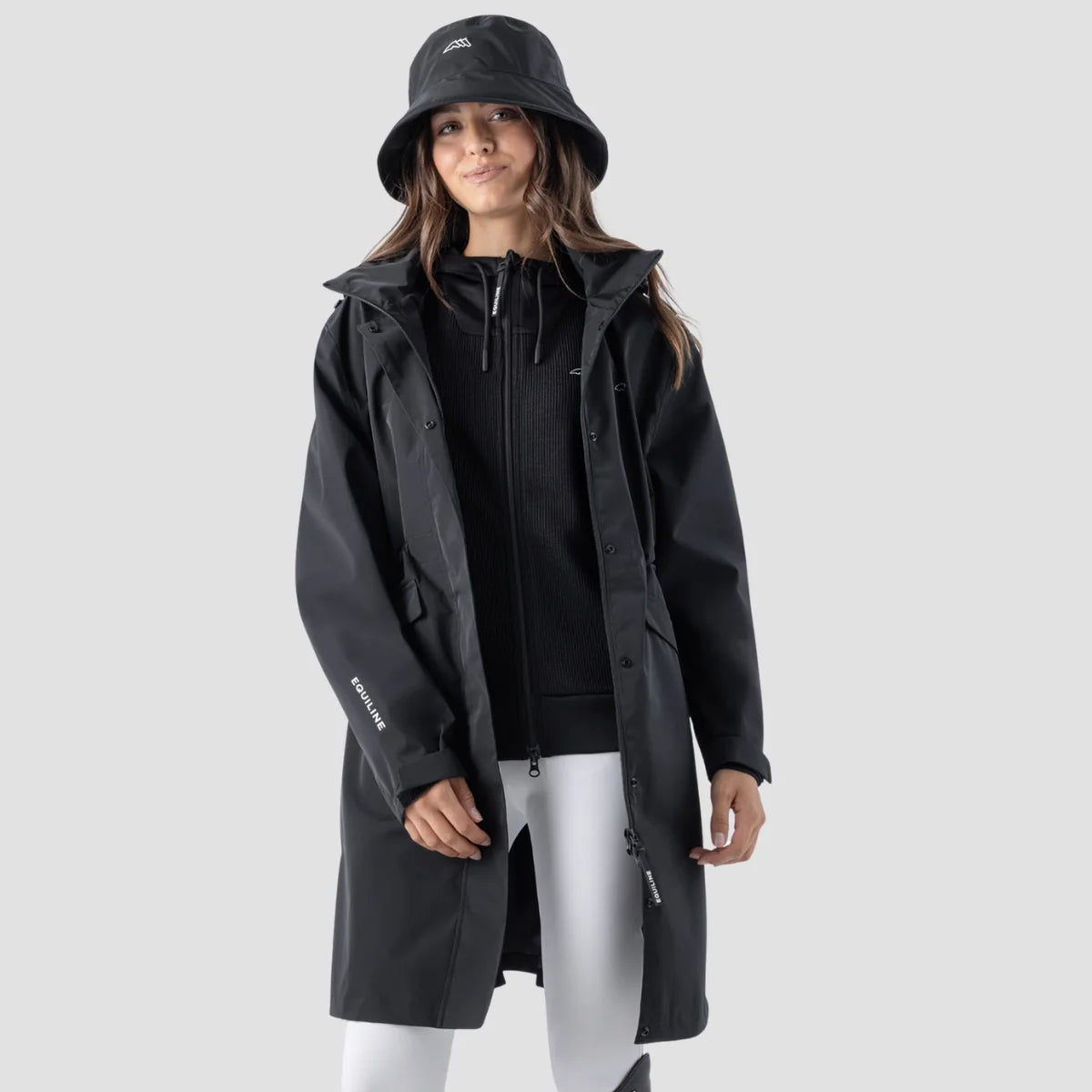 A woman modeling a black hooded raincoat against a light gray studio background. The coat has a small white logo on the left chest and a white logo on the sleeve.