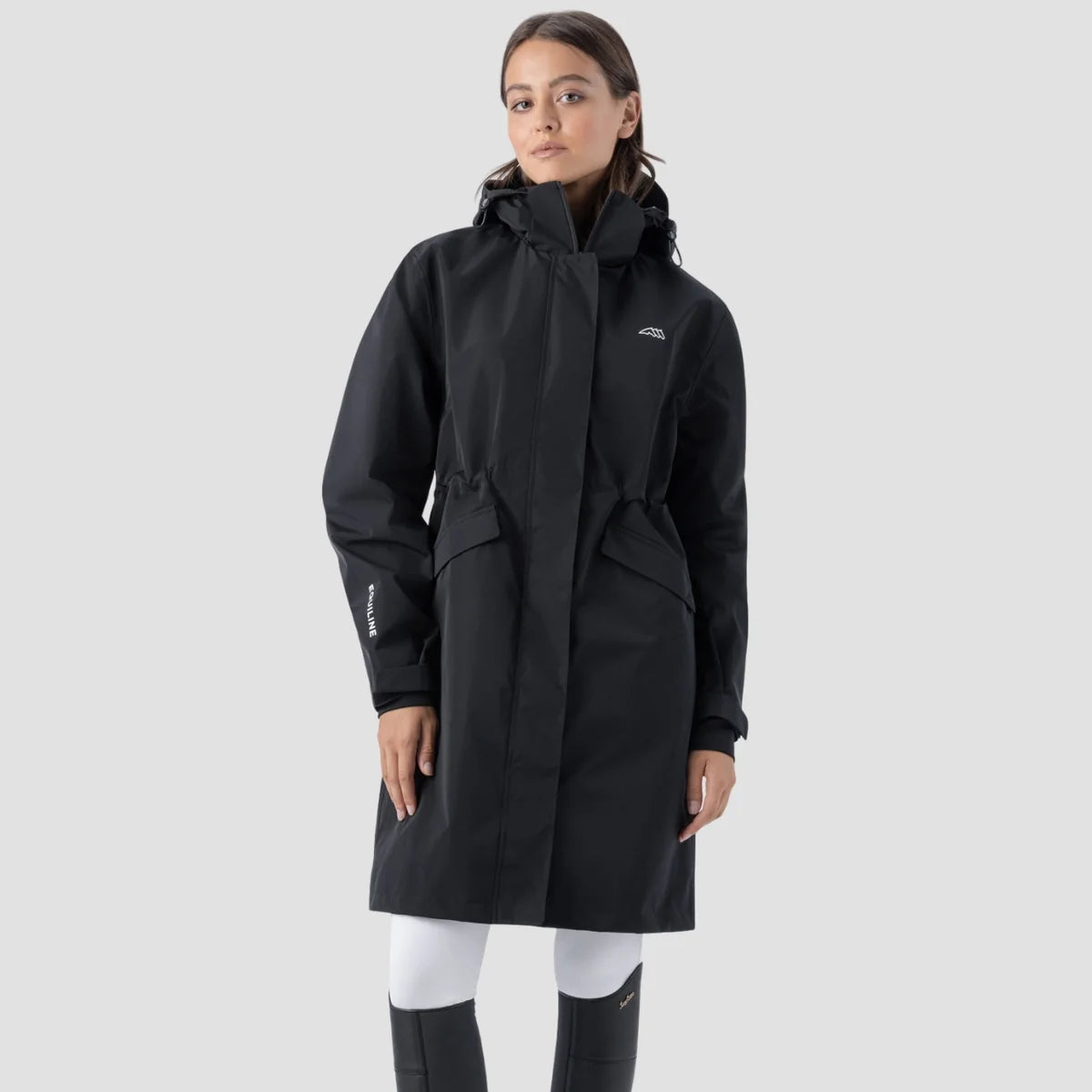 A woman modeling a black hooded raincoat against a light gray studio background. The coat has a small white logo on the left chest and a white logo on the sleeve.