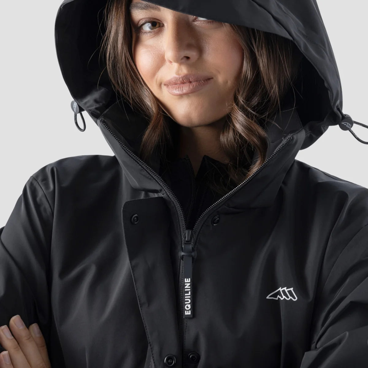 A close-up of a woman wearing a black hooded rain jacket with a front zipper, snap buttons, and a white Equiline logo on the chest, set against a plain light gray background.