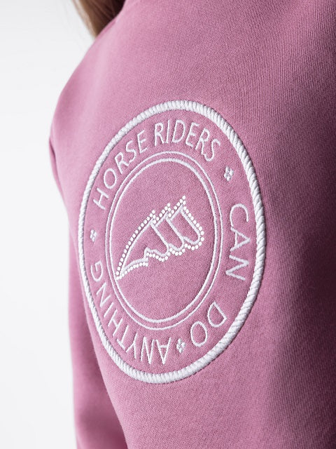 A close-up of a dusty pink sweatshirt featuring a circular white embroidered patch on the shoulder with the text 'HORSE RIDERS CAN DO ANYTHING' and a stylized horse rider emblem.