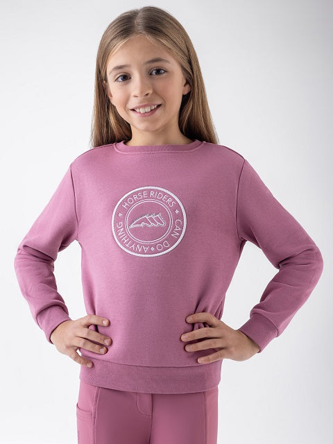 A young girl with long light brown hair smiling, wearing a dusty rose pink crewneck sweatshirt with a white circular 'Horse Riders Club Do Anything' graphic on the chest, paired with matching pink pants, against a light gray studio background.