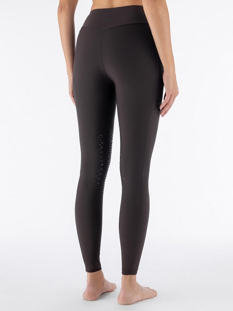 Rear view of a model wearing dark brown high-waisted leggings with textured grip dots on the outer side of one leg, standing barefoot against a white studio background.