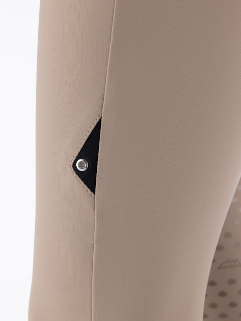 Close-up of beige leggings on a mannequin or model leg, showing a side seam and a small black triangular fabric detail with a silver snap button.