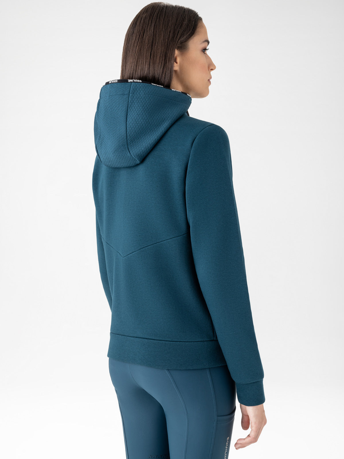 Woman wearing a teal full zip sweatshirt and riding tights on a white background