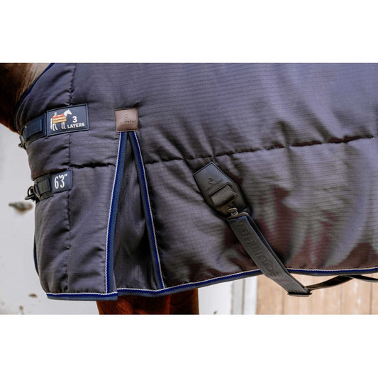 Close-up of a horse blanket with a dark gray textured body, blue piping, and black straps, shown on a horse.