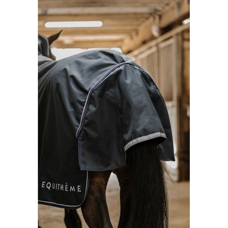 Horse wearing a dark gray raincoat with 'Equithème' branding in a stable.