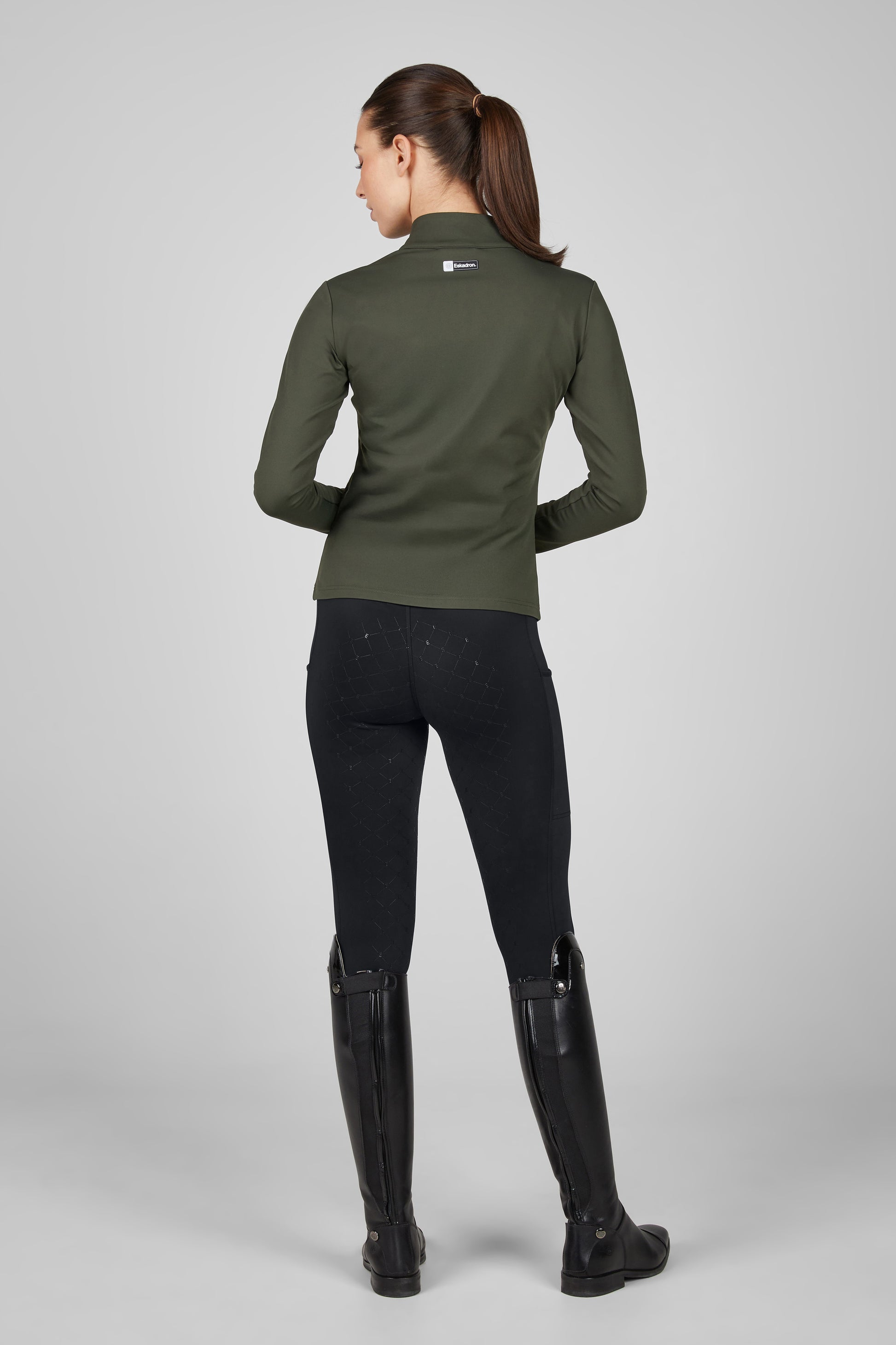 Person wearing a green equestrian jacket, black pants, and black riding boots on a gray background