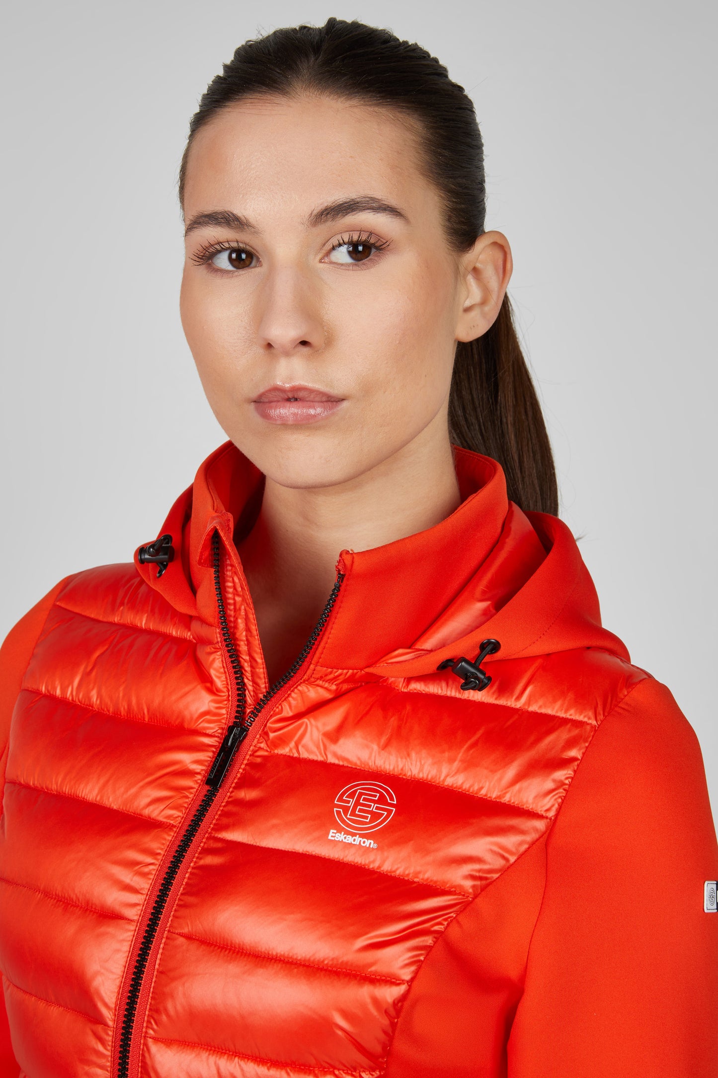Woman wearing an orange puffer jacket with a brand logo on a gray background