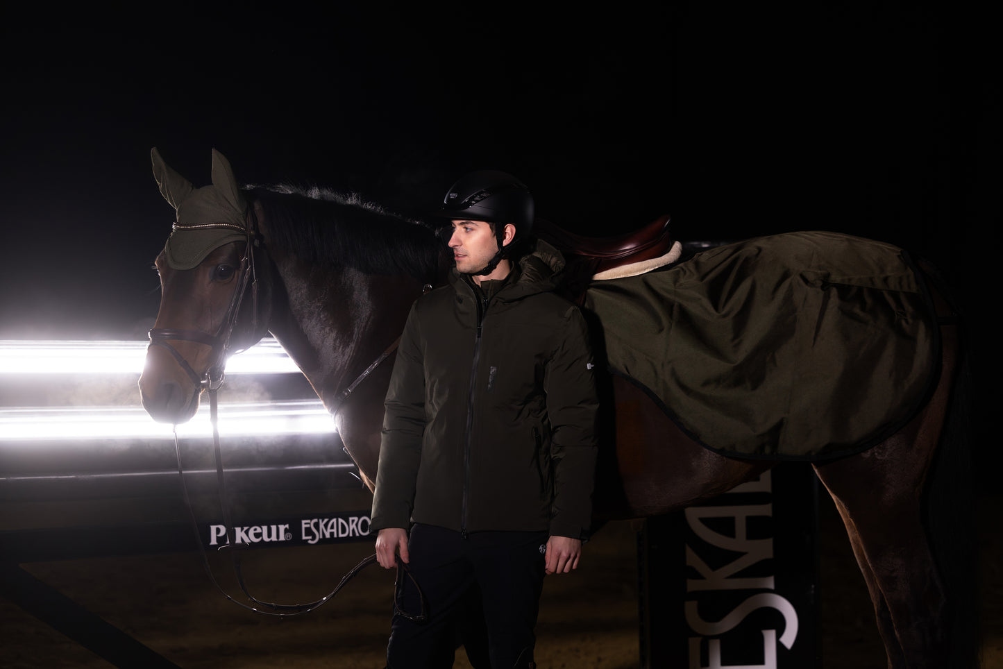 Person standing next to a horse in a dark setting with ESKADRON branding.