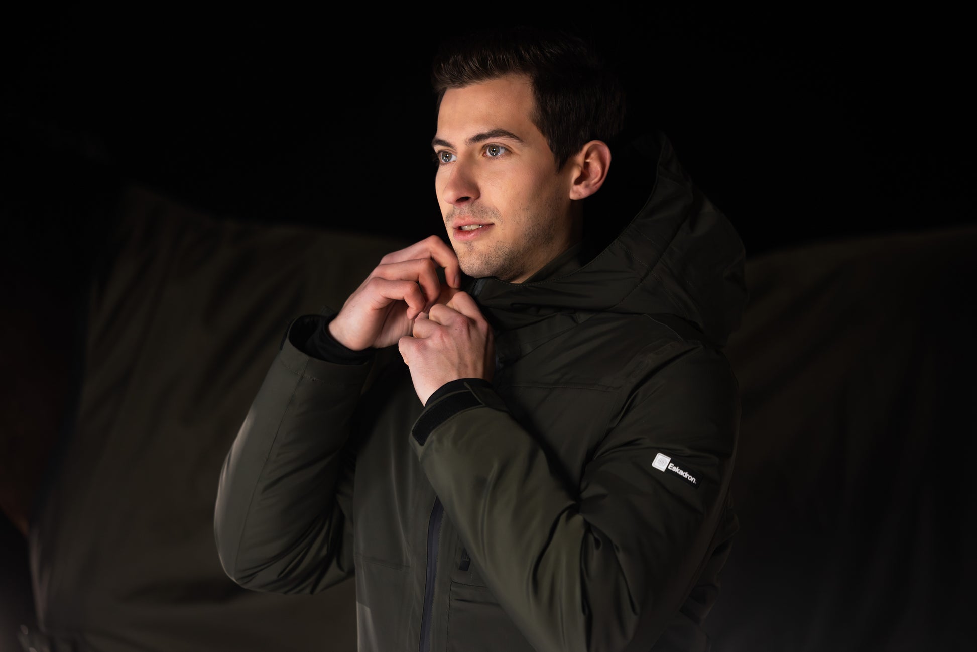 Man wearing a black jacket with a brand logo against a dark background