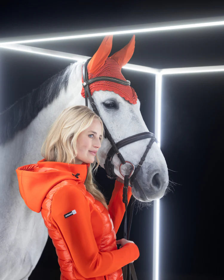 Woman in an orange jacket standing next to a white horse wearing an orange fly veil.