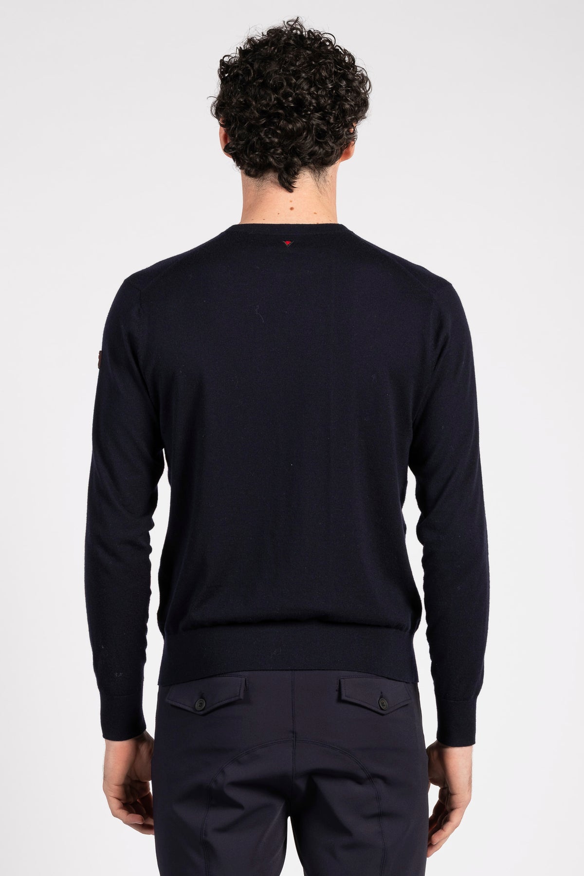 A man viewed from behind wearing a dark navy blue long-sleeve knit sweater and matching dark trousers against a light gray studio background.