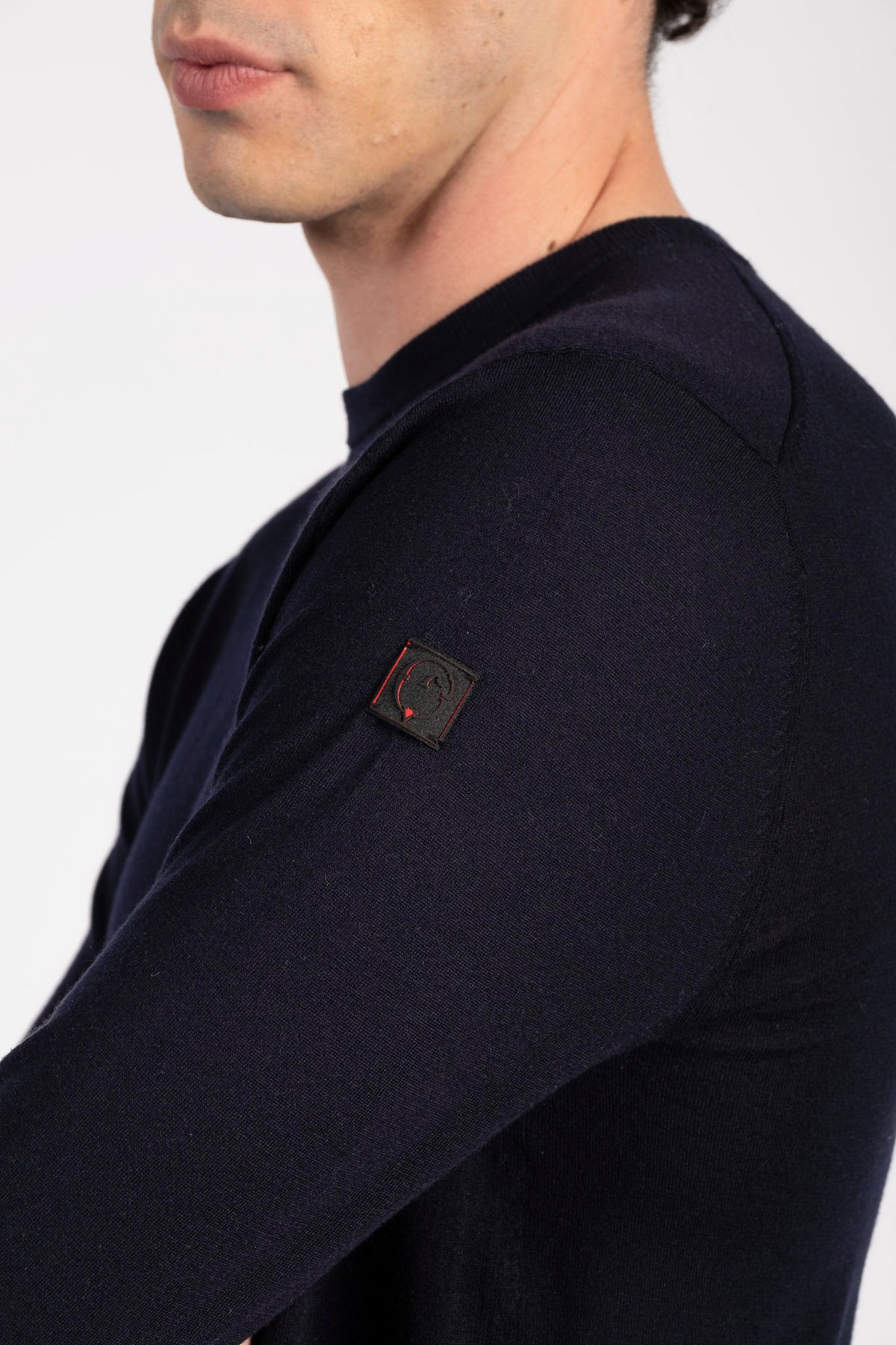 Close-up of a man wearing a dark navy long-sleeve knit top, showing a small square brand patch on the upper left shoulder.