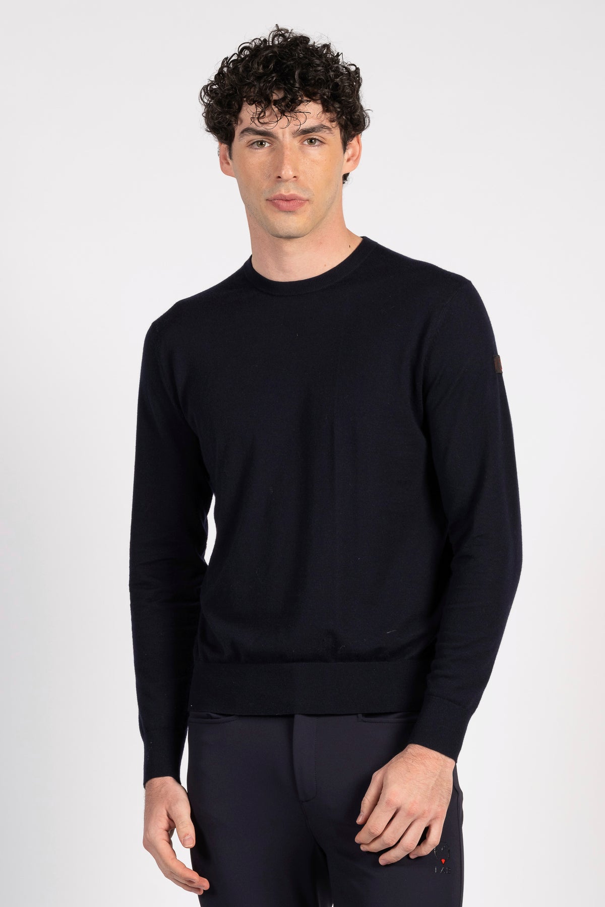 A man wearing a black long-sleeve crewneck sweater and matching black trousers, standing against a light gray studio background.