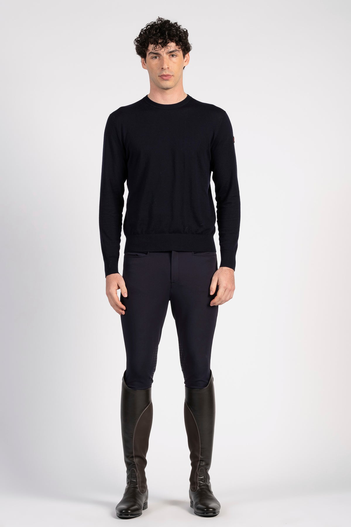 A man modeling a black long-sleeve crewneck sweater, dark navy riding breeches, and black riding boots against a light gray studio background.