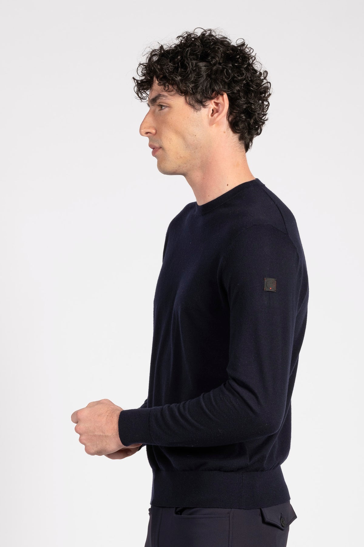 A man in profile wearing a dark navy long-sleeve knit sweater with a small square logo patch on the upper left sleeve, photographed against a clean light gray studio background.