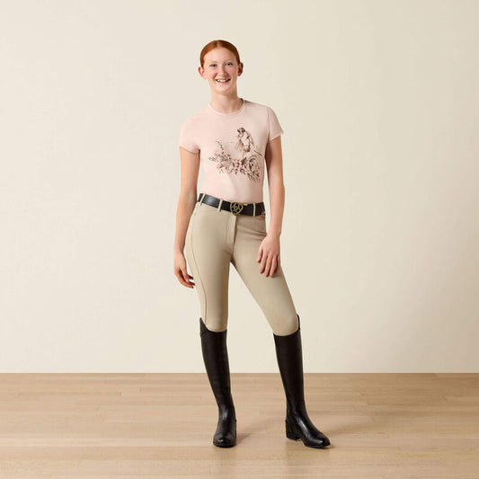 Woman wearing a pink shirt with floral design, beige riding pants, and black riding boots on a plain background.