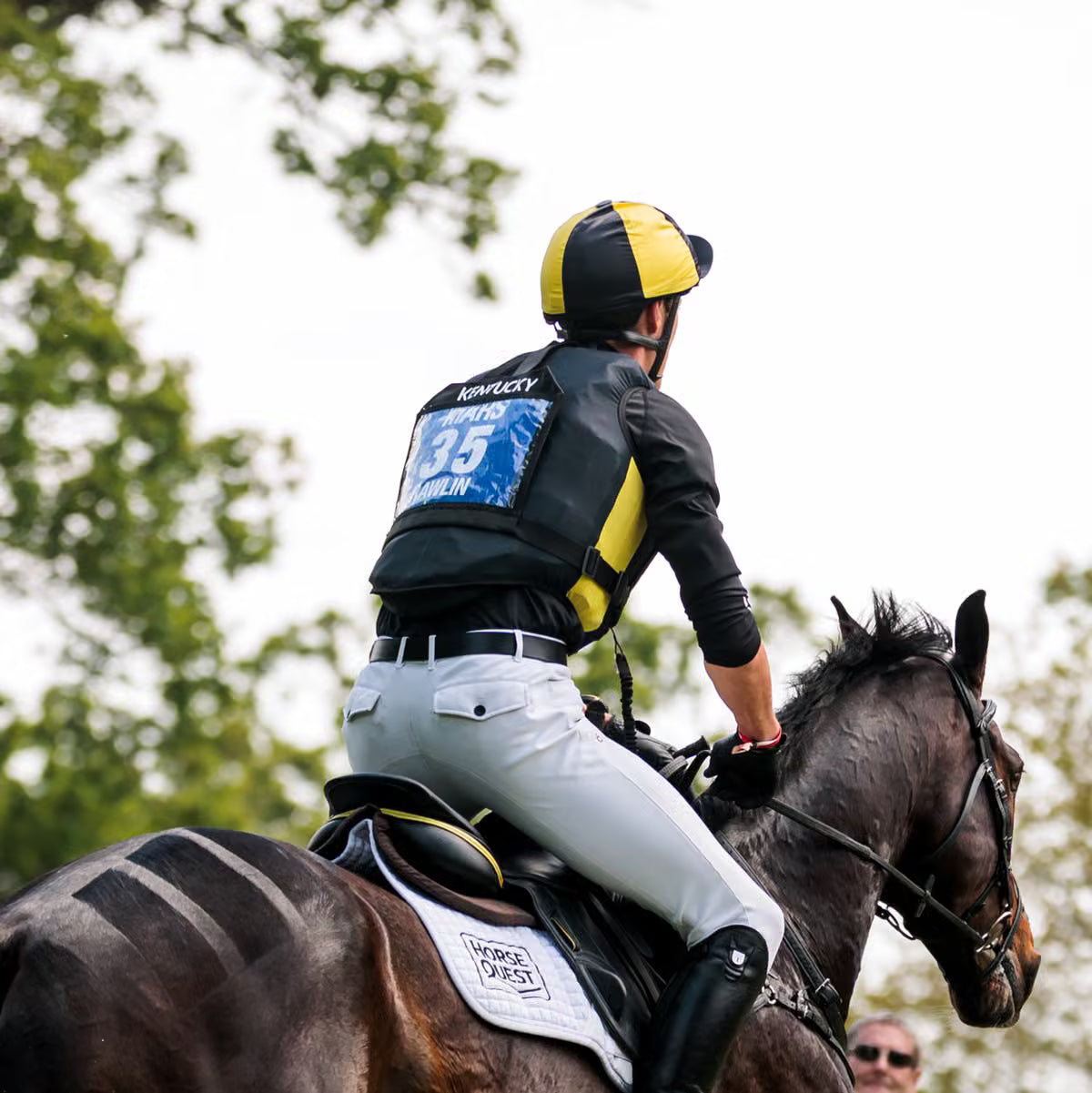 A jockey wearing a black and yellow helmet and matching black and yellow vest with white pants rides a dark brown horse outdoors. The jockey's back is turned, showing the number 35 on the back.