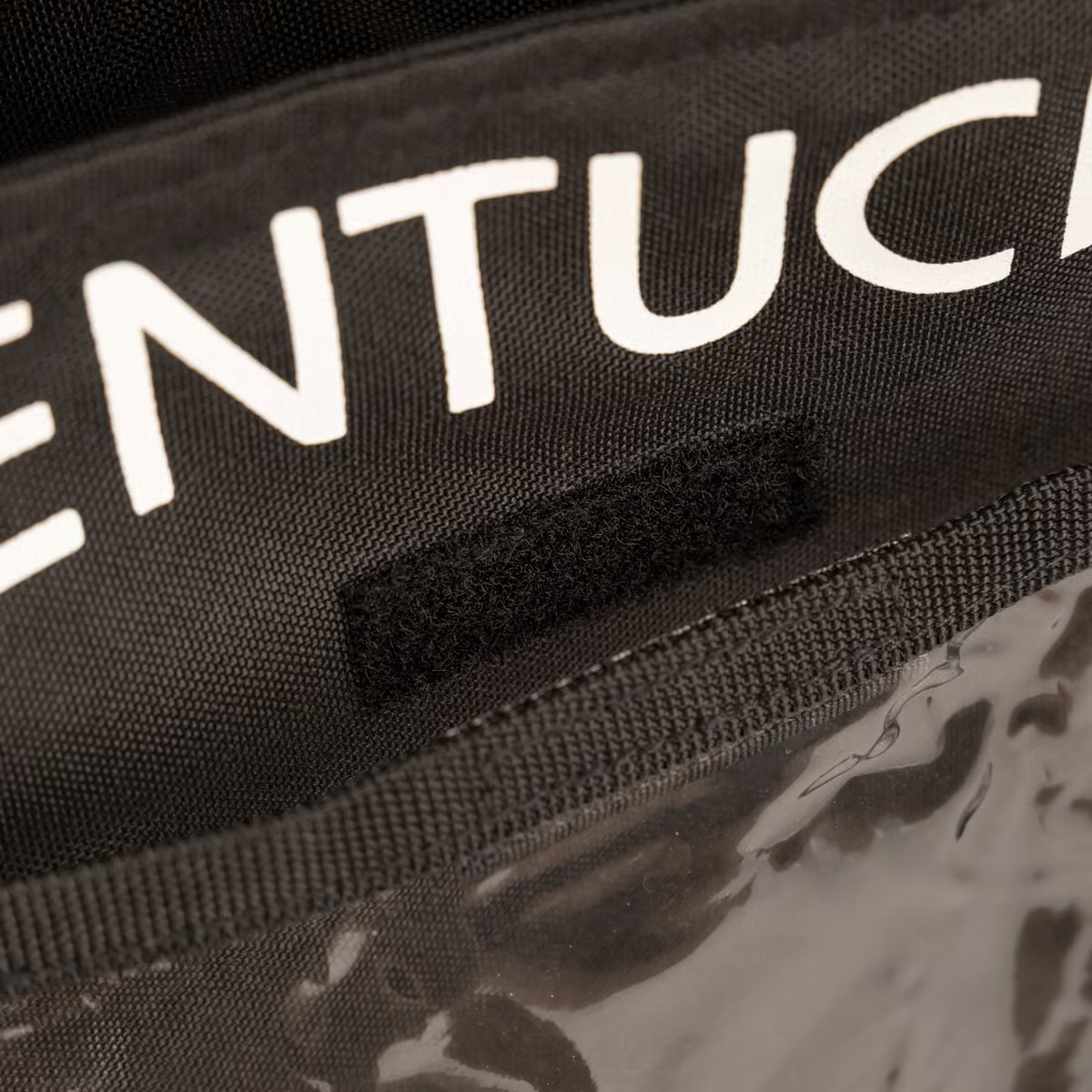 Close-up of a black fabric bag with white partial lettering reading 'KENTUCKY' and a black hook-and-loop strap, above a clear plastic panel.
