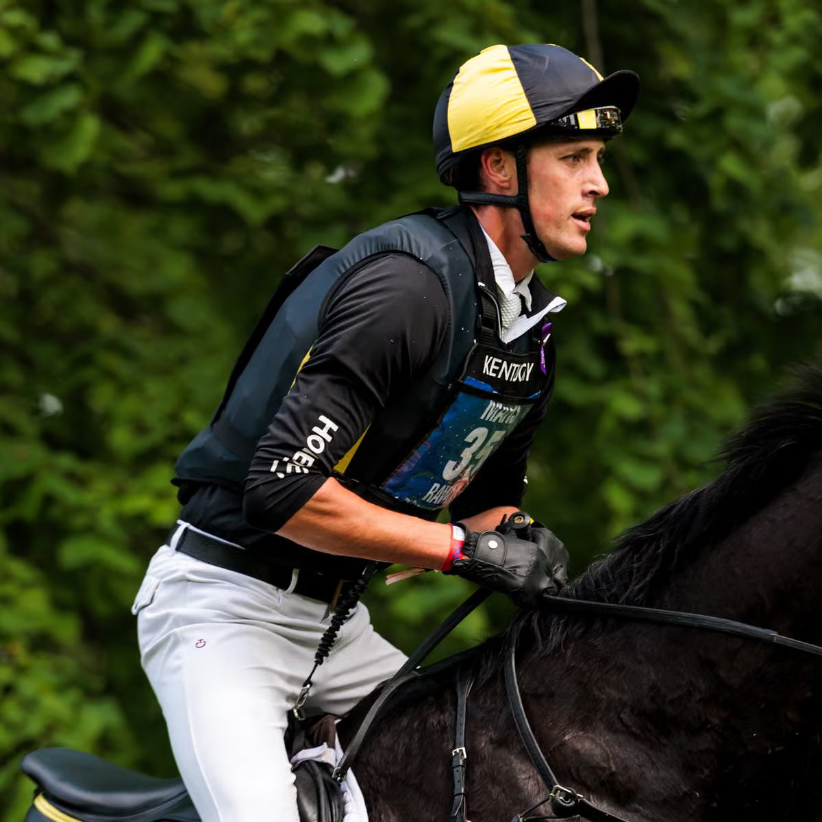 A male jockey riding a dark horse outdoors, wearing a black and yellow helmet, a black jersey with 'KENTUCKY' and number 35, white pants, and black gloves. The background is a dense, out-of-focus green forest.