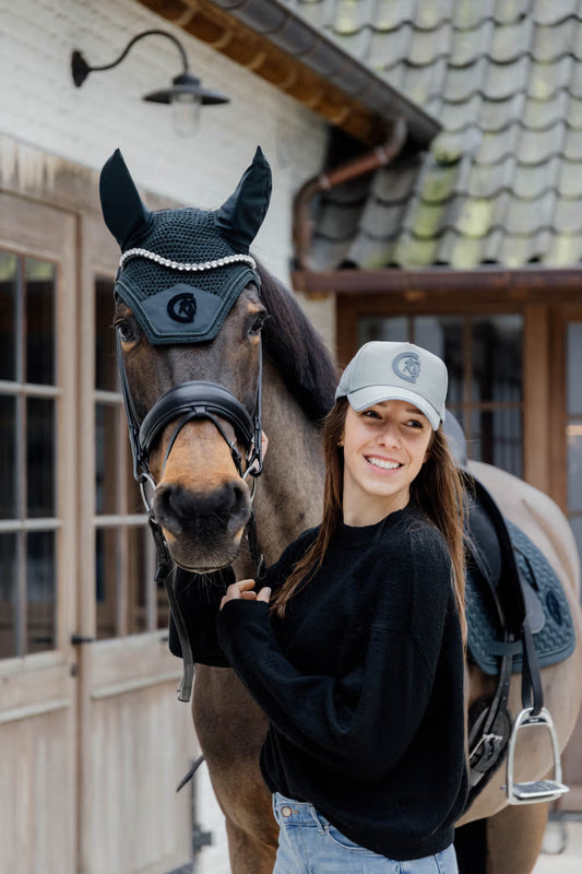 Woman standing next to a horse wearing a bridle and a fly veil in front of a wooden building.