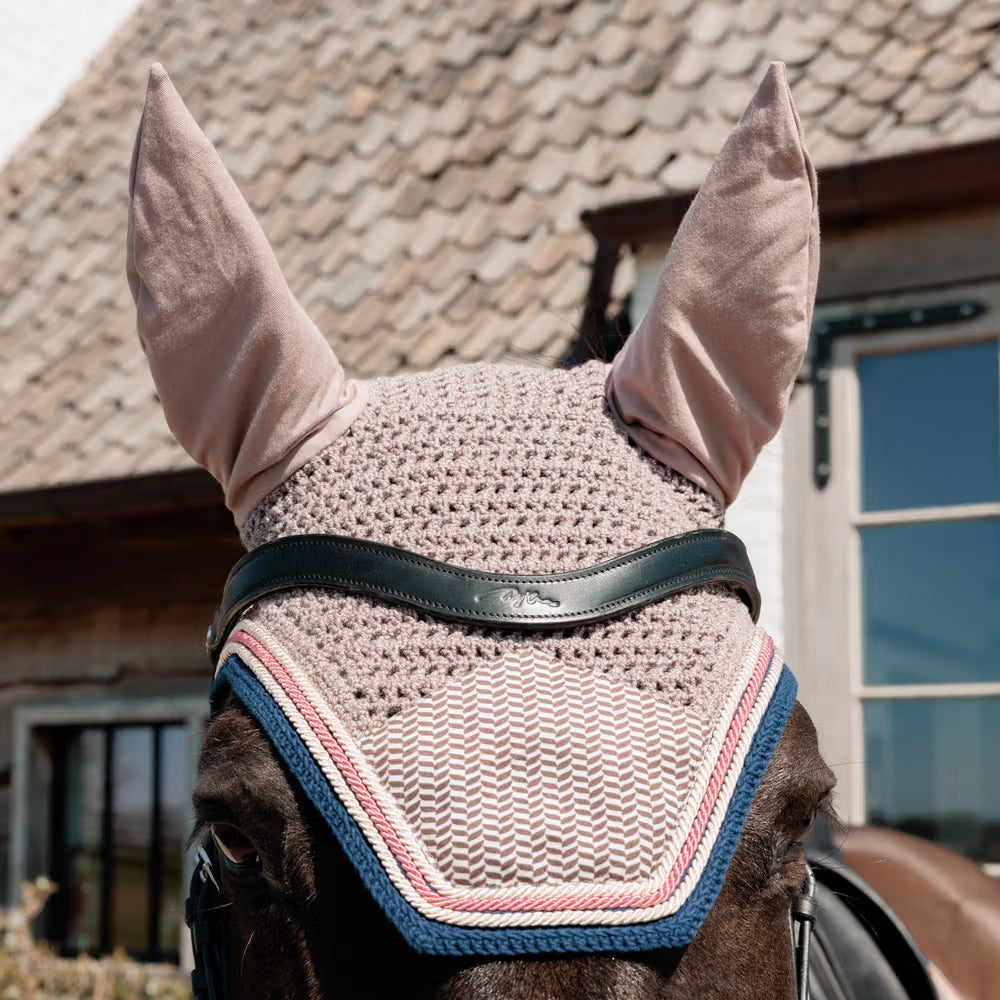 Horse wearing a beige fly mask with pink earflaps in front of a building.