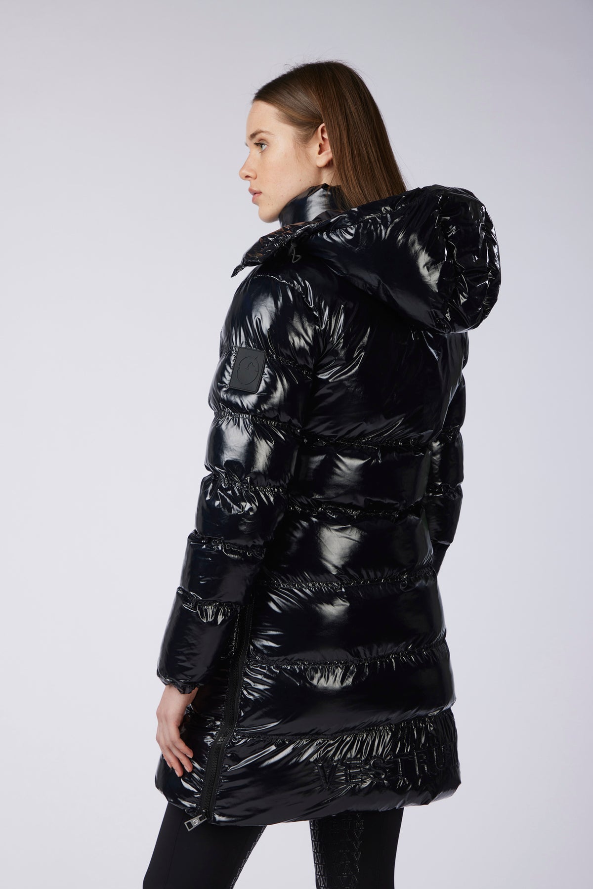 A woman wearing a shiny black puffer jacket with a high shiny hood, shown from a side-back angle against a light gray studio background.