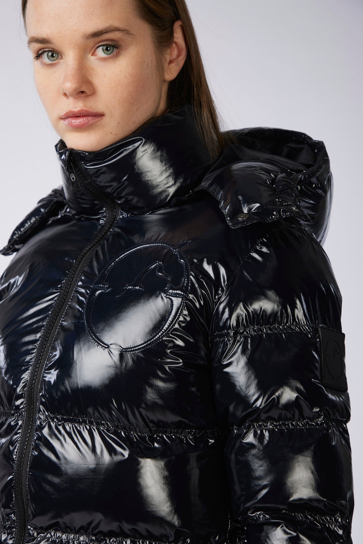 A close-up of a model wearing a shiny black puffer jacket with a high collar and quilted horizontal stitching against a light gray background.