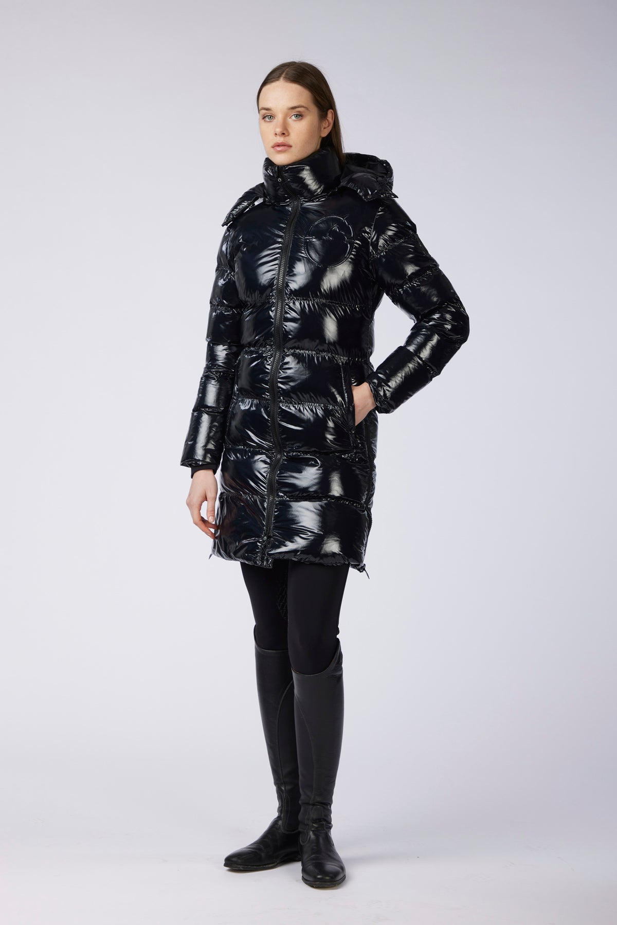 A woman modeling a shiny black puffer coat with a high collar and hood, paired with black leggings and black boots, against a light gray studio background.